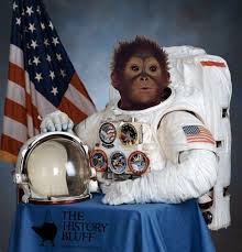Gordo posing in space suit with his helmet off looking to the side with an American flag in the background.