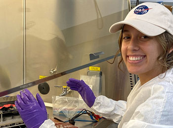 smiling female lab tech wearing nasa hat working on substances.